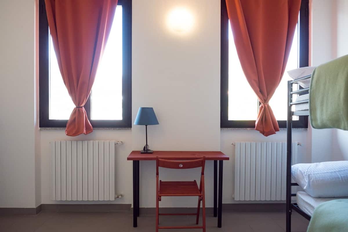 A room with a table, a chair, a lamp, two windows with curtains, a radiator, and a bunk bed.