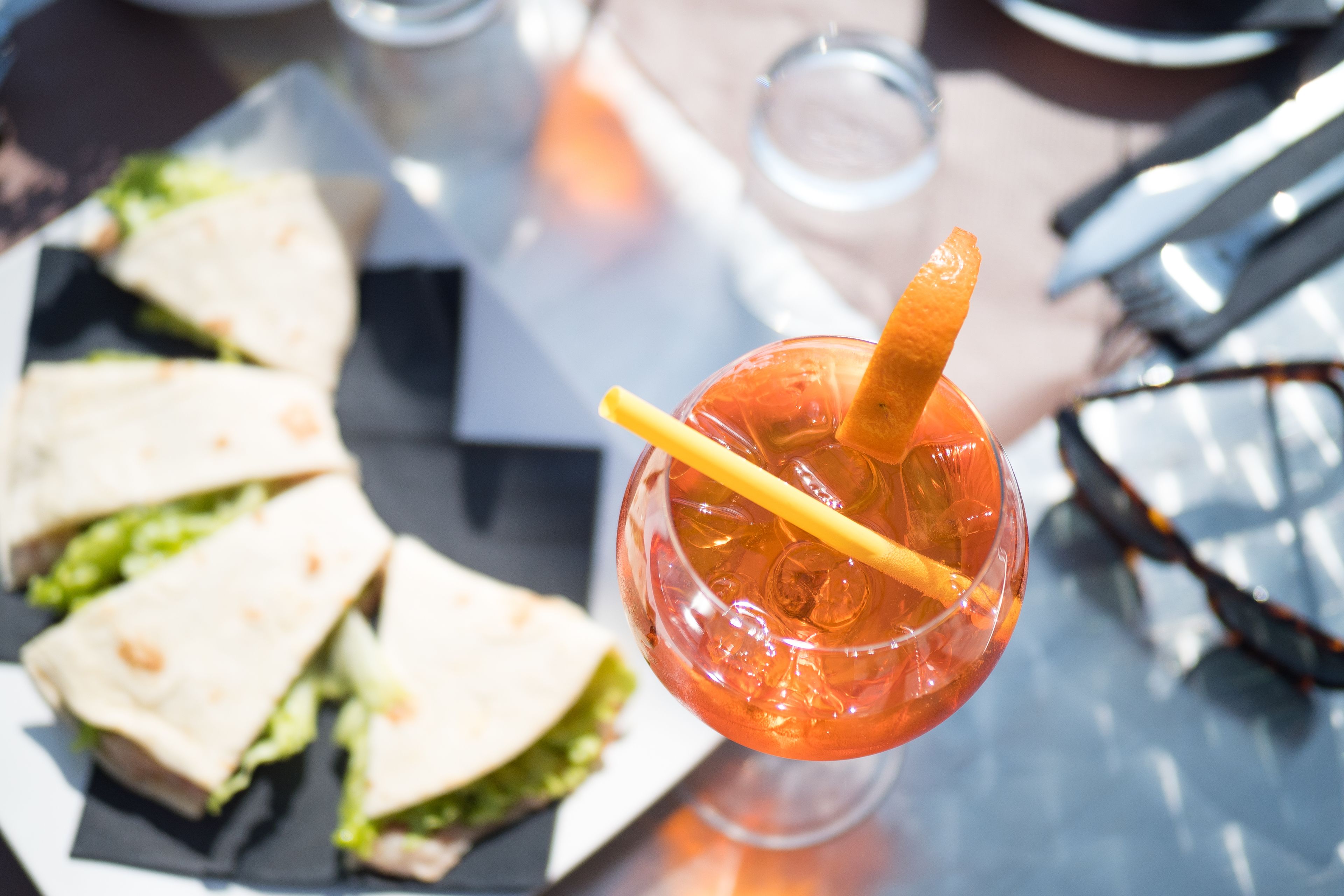 A glass of iced drink with a straw and a slice of orange, accompanied by a plate of sandwiches.