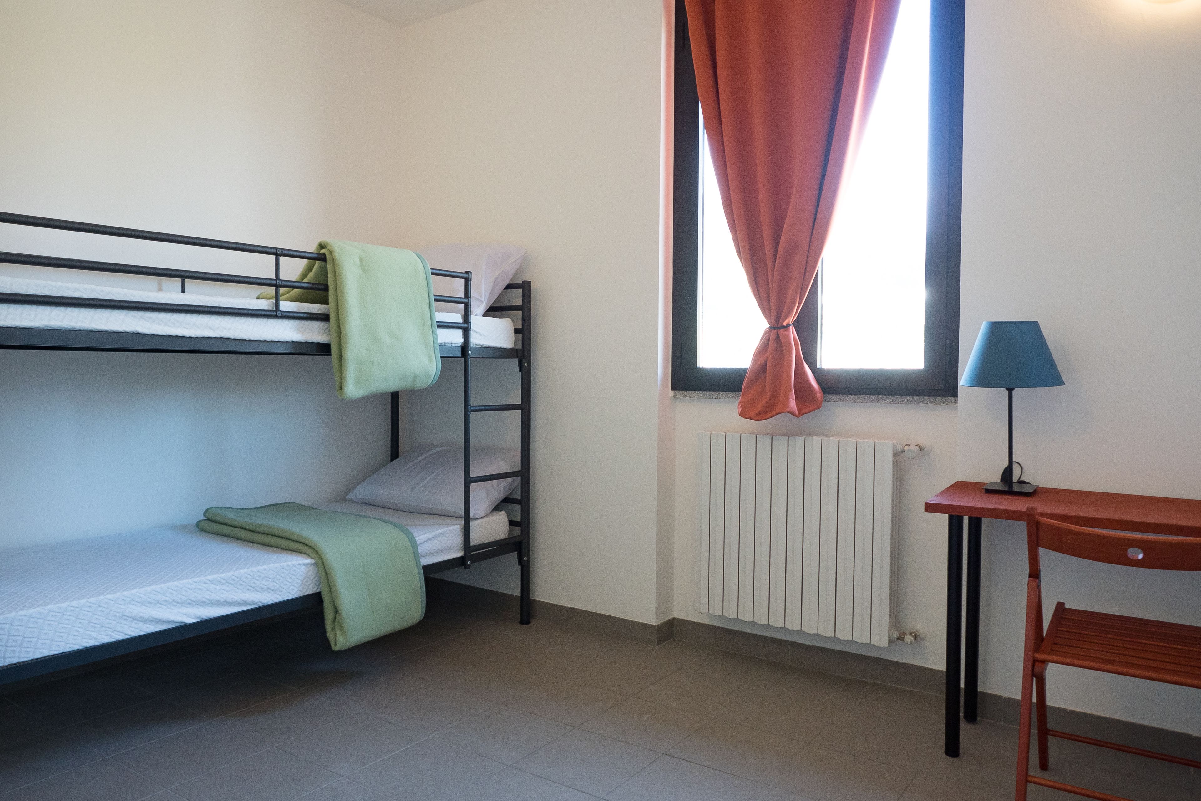 A dormitory room with bunk beds, a desk, a chair, a lamp, and a window with curtains.