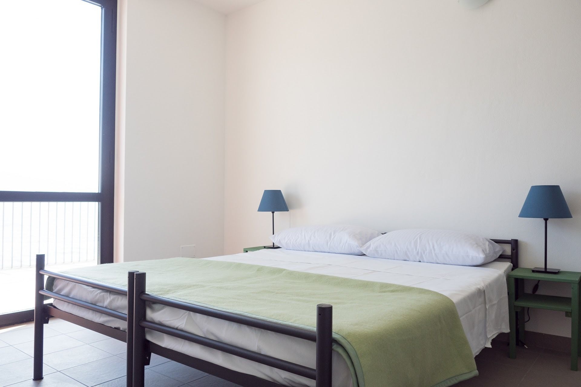 Ein Einzelbett mit einer grünen Decke, zwei Nachttische mit Lampen und ein Fenster mit Jalousien.