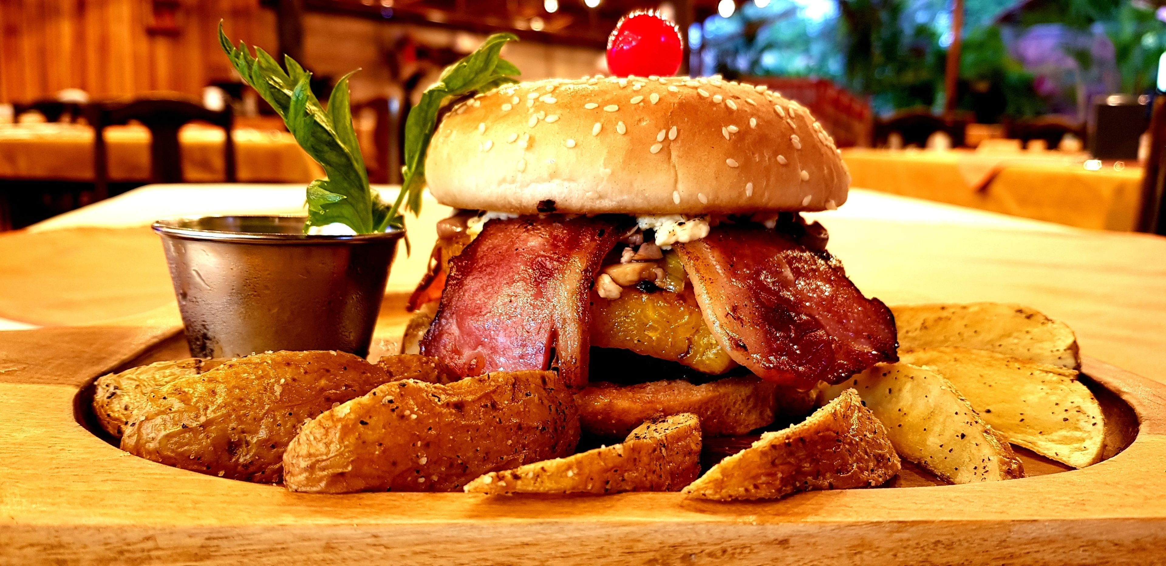 Ein Burger mit Speck, Salat und einer Kirsche, serviert mit Bratkartoffeln auf einer Holzplatte.