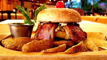 Ein Burger mit Speck, Salat und einer Kirsche, serviert mit Bratkartoffeln auf einer Holzplatte.