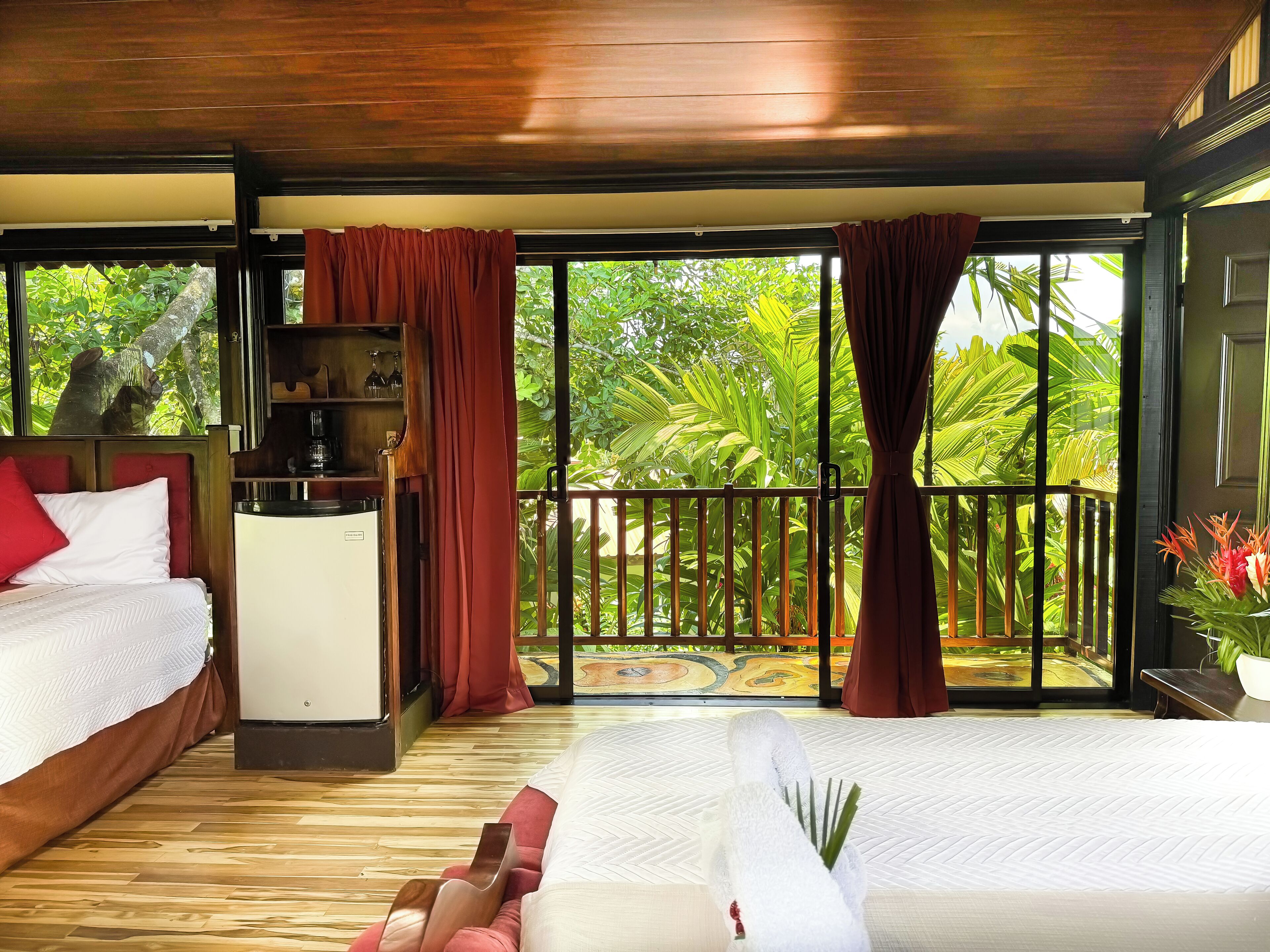 A room with two beds, a wooden ceiling, a small desk, and a view of lush greenery through large windows.