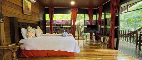 A wooden-floored bedroom with a bed, a TV, and a view of greenery outside.
