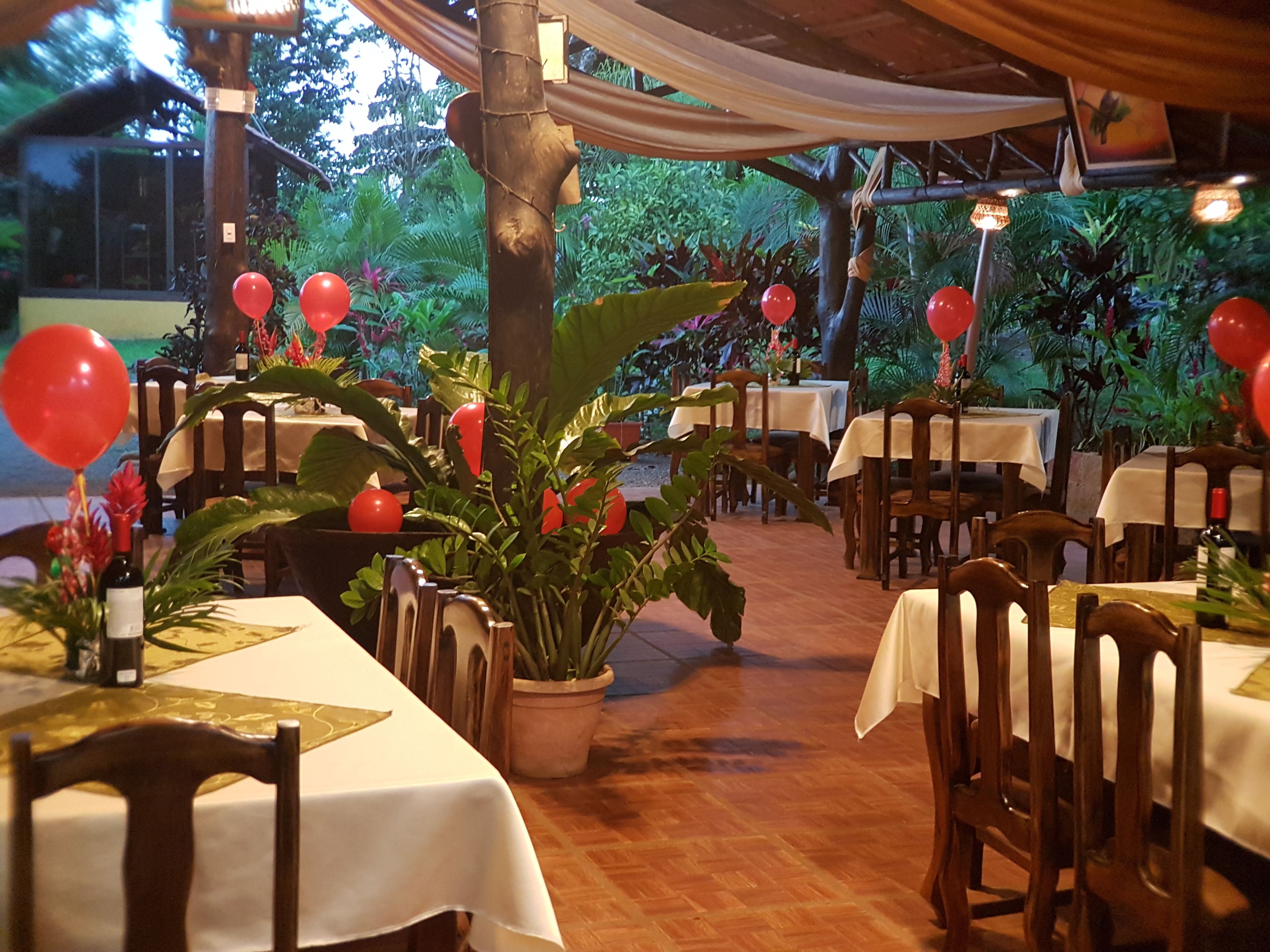 An outdoor dining area with tables and chairs, surrounded by potted plants and red balloons.