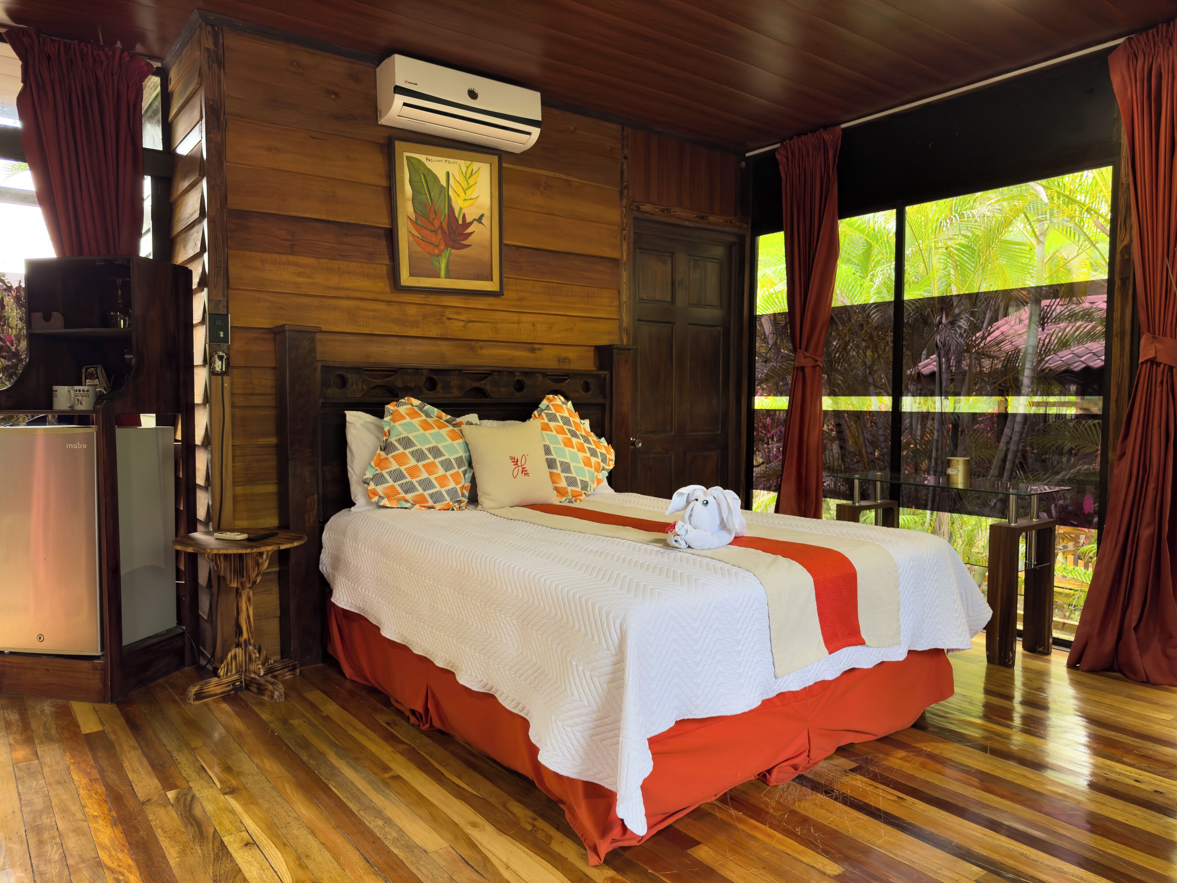 A wooden room with a bed, an air conditioner, a window with curtains, and a view of greenery outside.