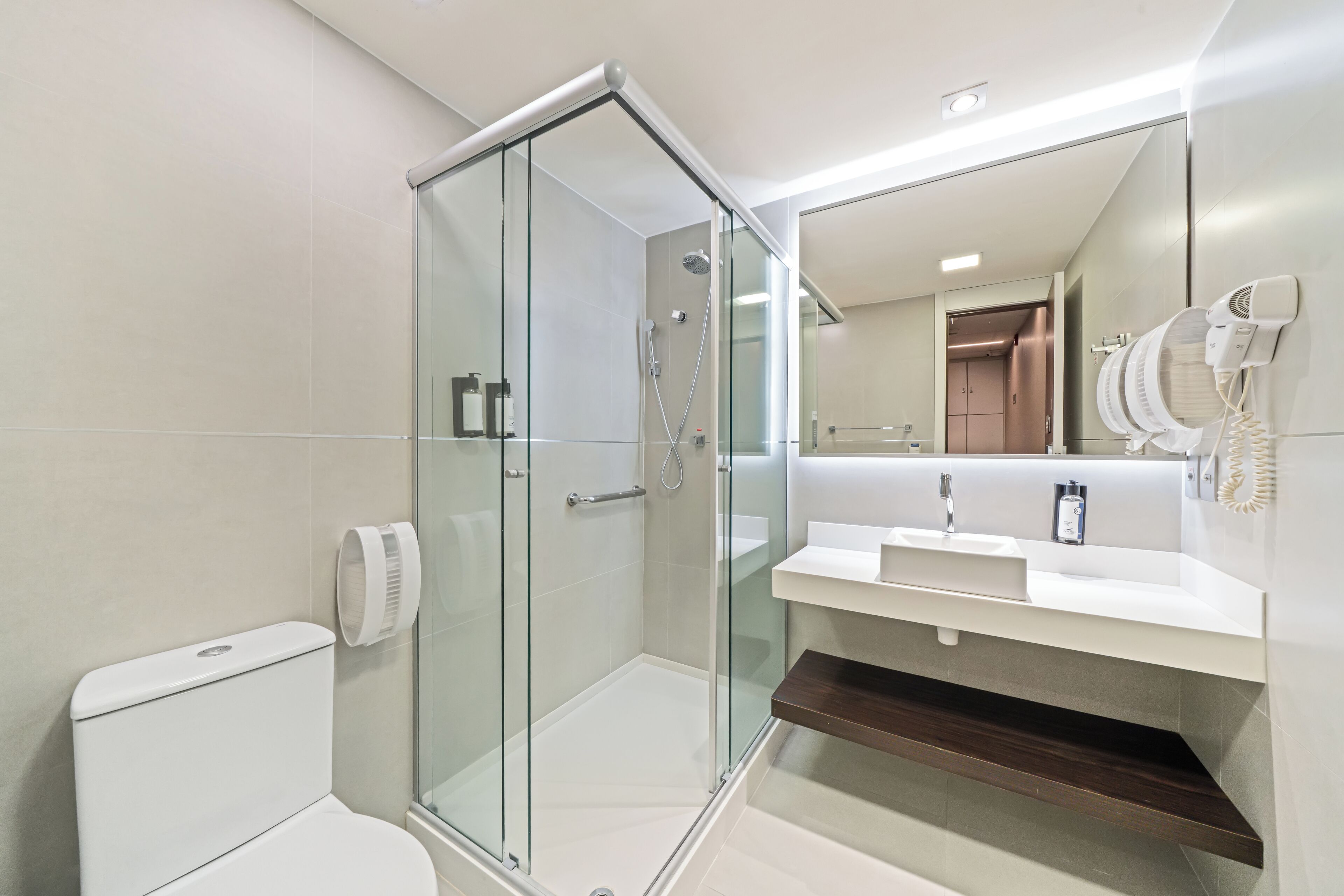 Un baño moderno con ducha de vidrio, inodoro blanco, lavamanos con espejo y secador de pelo montado en la pared.