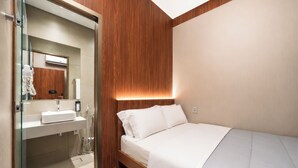 Une chambre d’hôtel moderne dotée d’une douche en verre, d’un mur d’accent en bois, d’un lit avec une literie blanche et d’un appareil de climatisation visible.