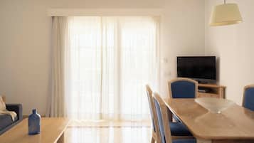Una sala de estar con una mesa de comedor de madera, sillas tapizadas en azul, un sofá, un televisor sobre un mueble bajo y un ventanal con cortinas de encaje.