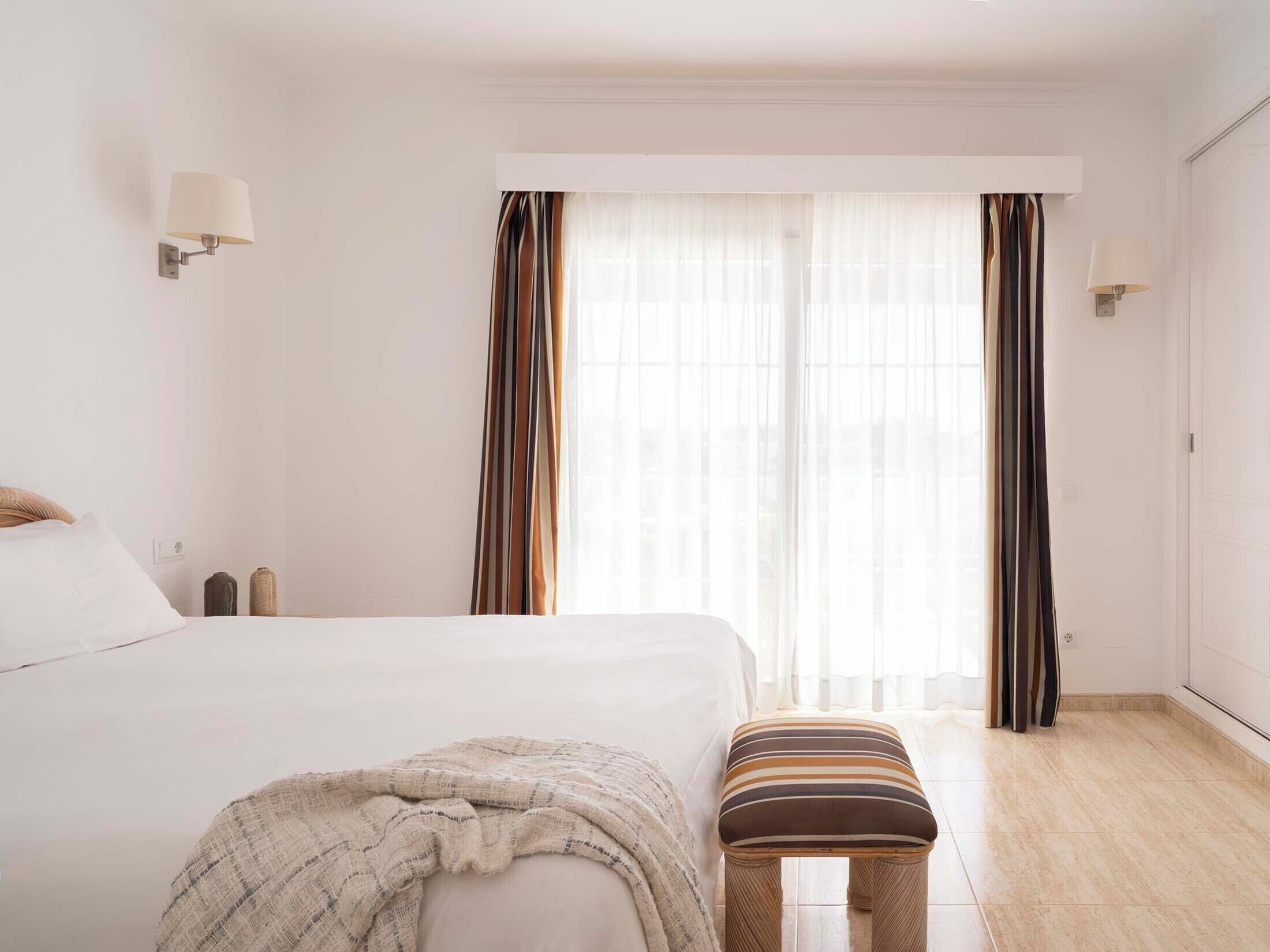 Quarto de hotel com cama, um banco, janela com cortinas e luminárias fixadas na parede.