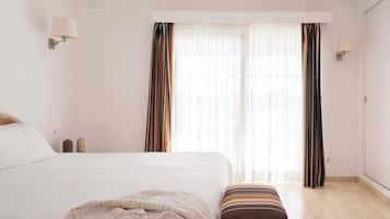 Quarto de hotel com cama, um banco, janela com cortinas e luminárias fixadas na parede.