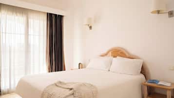 Habitación de hotel con una cama grande, una mesita de noche con lámpara y vistas al exterior a través de cortinas translúcidas.