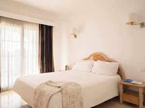 Une chambre d’hôtel avec un grand lit, une table de chevet avec une lampe et une vue sur l’extérieur à travers des rideaux légers.