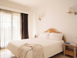 Habitación de hotel con una cama grande, una mesita de noche con lámpara y vistas al exterior a través de cortinas translúcidas.