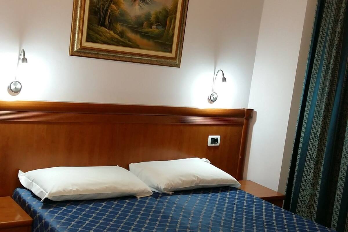 A hotel room with a wooden headboard, a bed with a blue checkered bedspread, two white pillows, and a painting of a landscape on the wall.