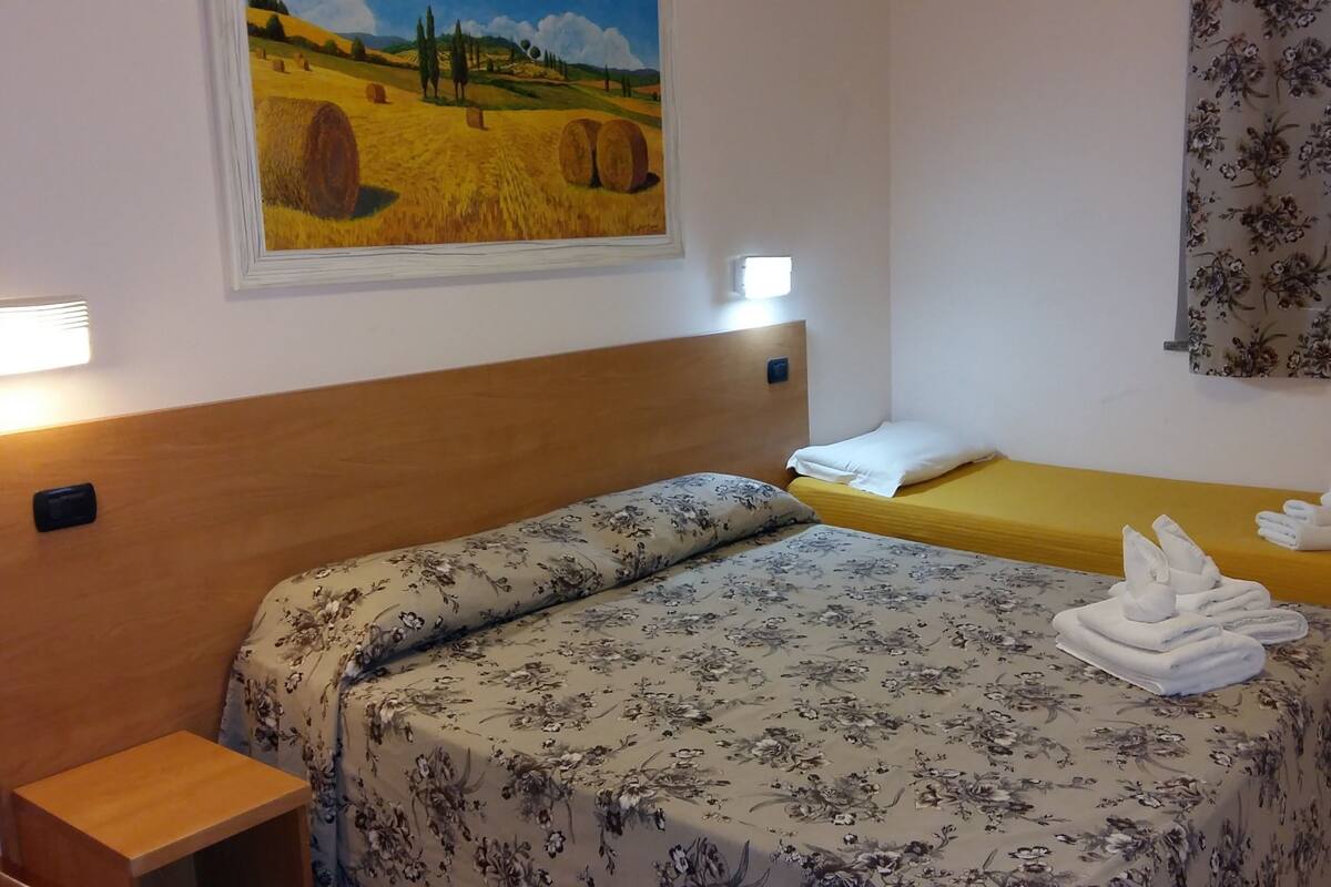 A hotel room with a wooden headboard, two beds with floral bedding, a painting of a field, and a small bedside table.
