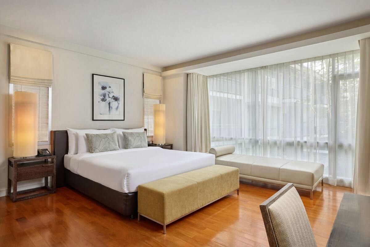 Habitación de hotel con una cama grande, lámparas de noche, un banco, un sofá y una ventana con cortinas translúcidas.
