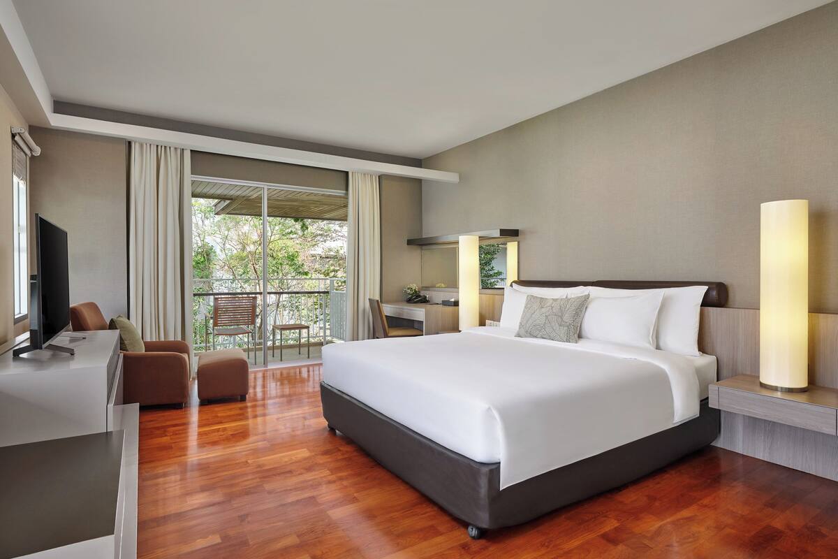 Habitación de hotel con cama, escritorio, silla, televisión, balcón con cortinas y lámpara.