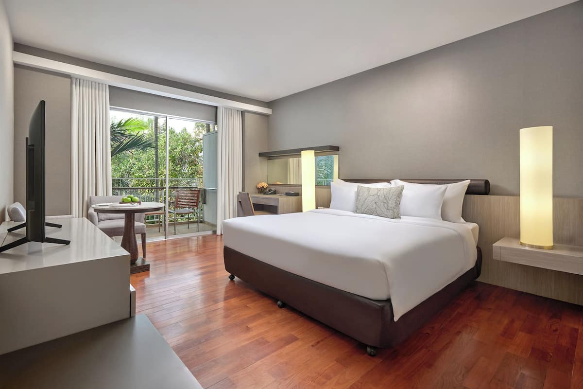 Habitación de hotel moderna con una cama grande, un escritorio, una silla, un televisor, un balcón con vistas a zonas verdes y una mesita de noche con lámpara.