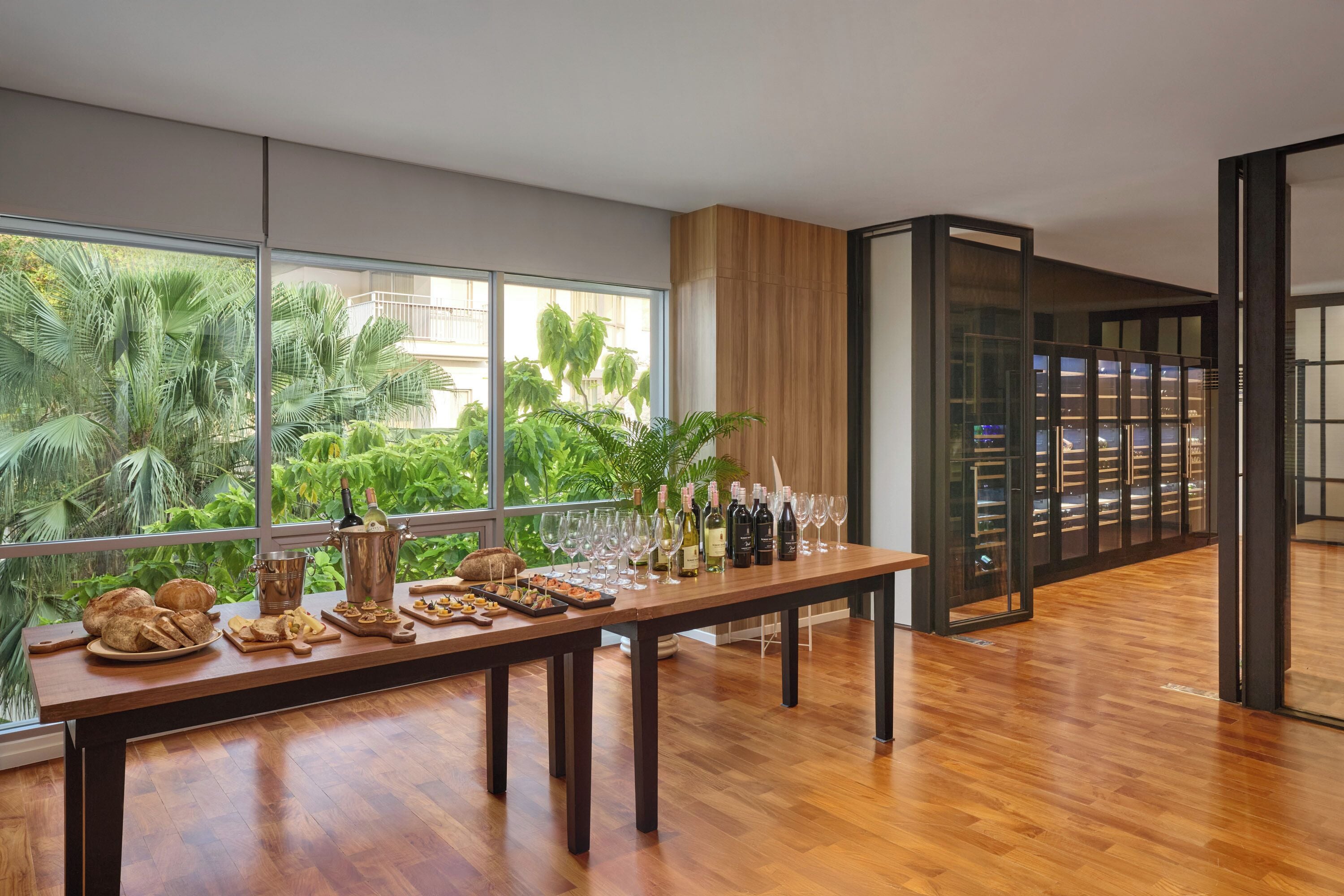 Ein geräumiges Zimmer mit einem langen Holztisch, der mit Speisen und Wein gedeckt ist, großen Fenstern mit Blick auf viel Grün und einem modernen Interieur.