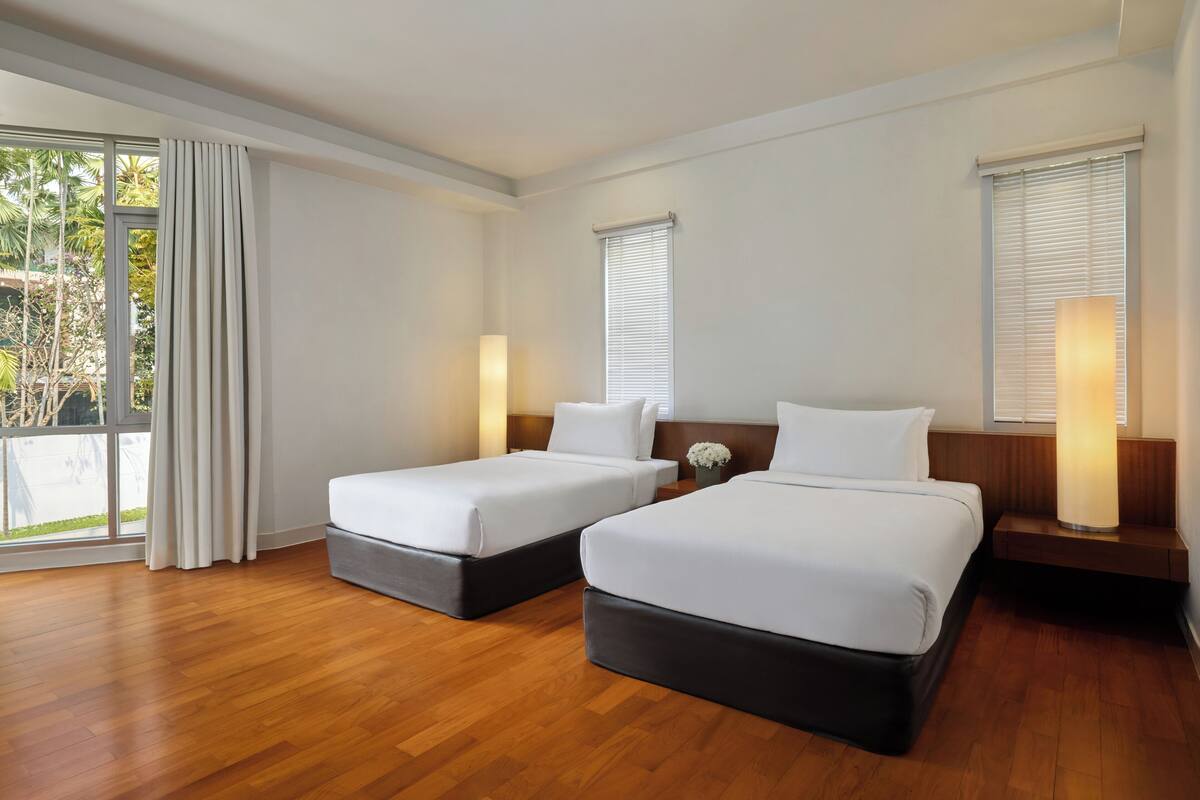 Habitación de hotel con dos camas, suelo de madera, un ventanal grande con cortinas y una lámpara fijada a la pared.