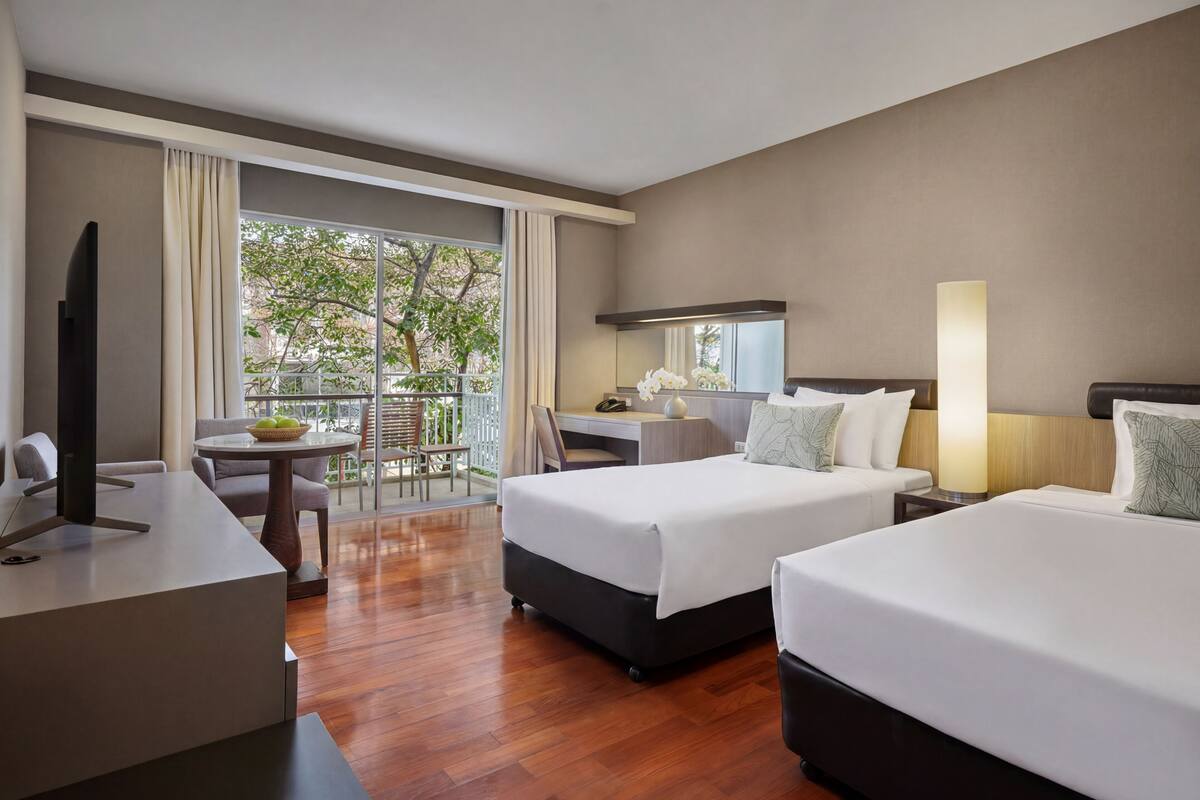 Habitación de hotel con dos camas, un escritorio, una silla, televisión y balcón con vistas a árboles.