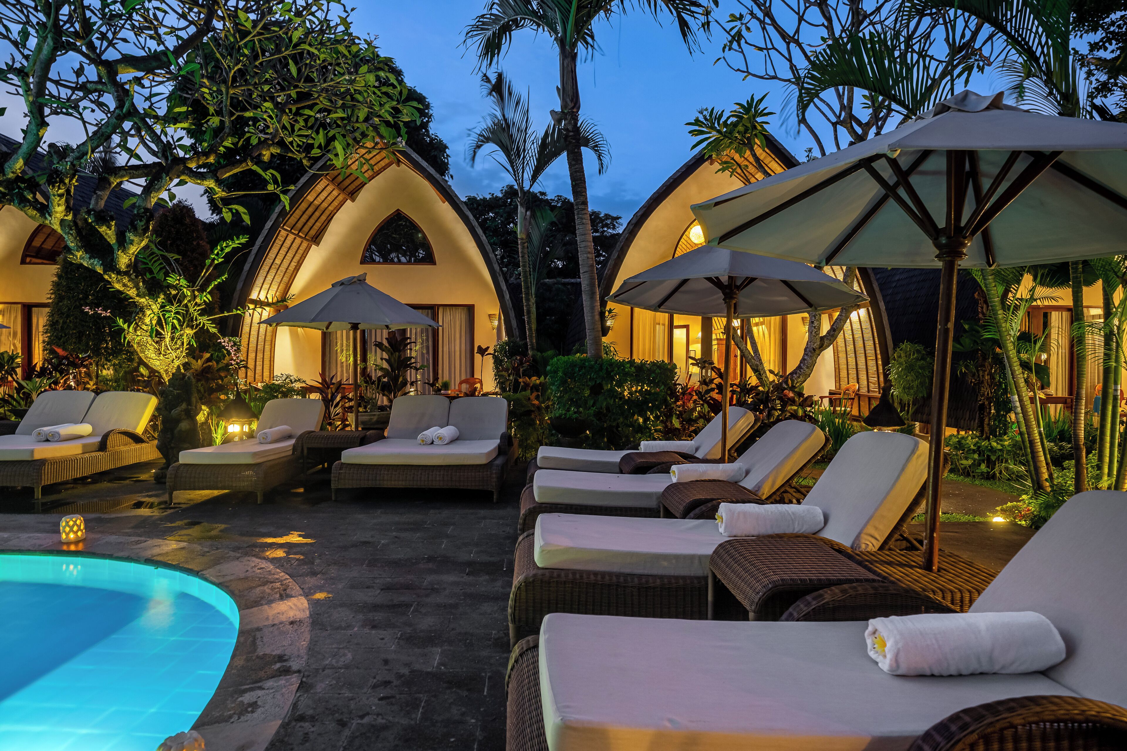 A poolside area with lounge chairs and umbrellas, surrounded by lush greenery and a clear pool.