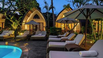A poolside area with lounge chairs and umbrellas, surrounded by lush greenery and a clear pool.