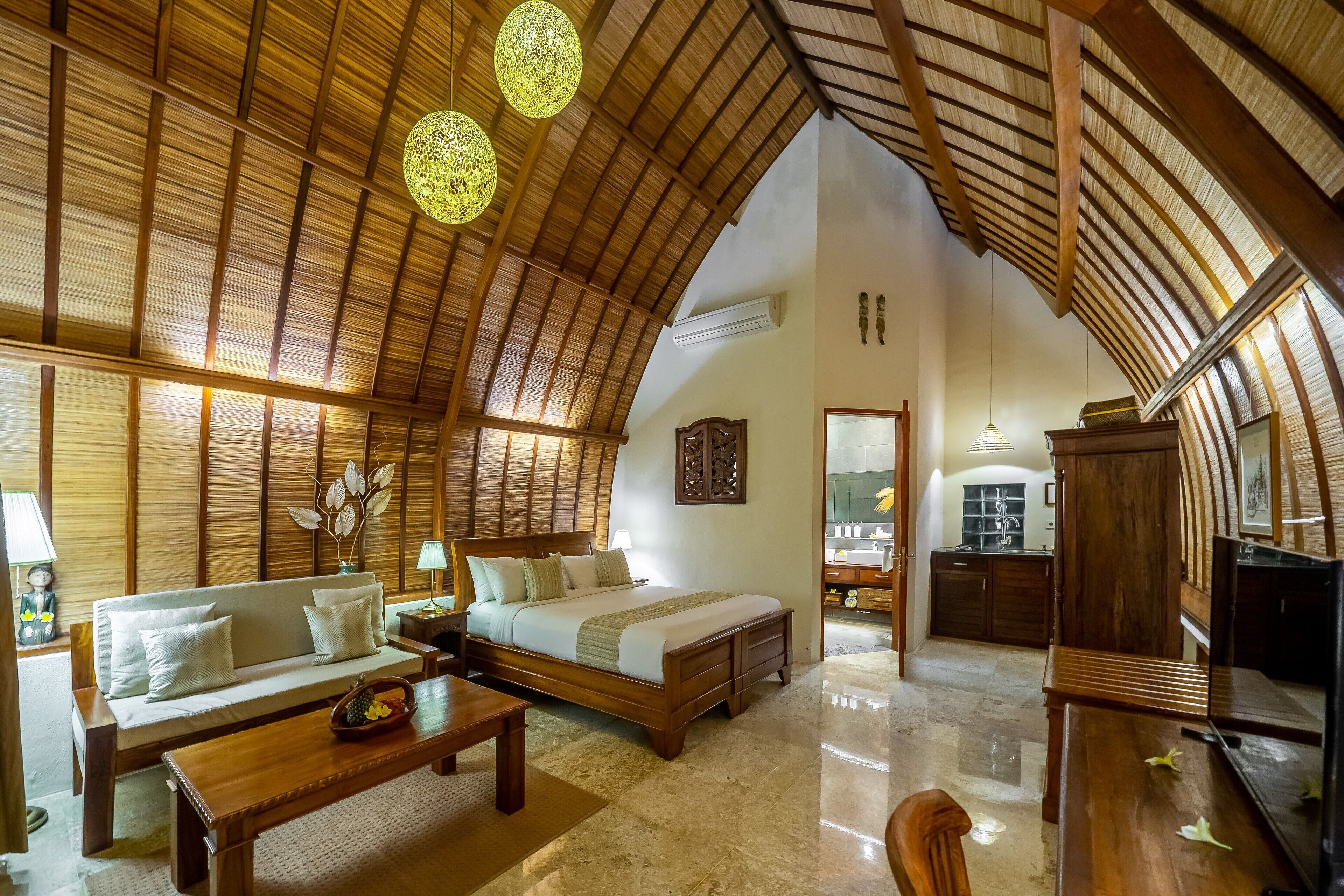 A spacious room with a high, sloped ceiling and wooden beams, a large bed with a wooden headboard, a sofa, a wooden coffee table, and a small dining table.
