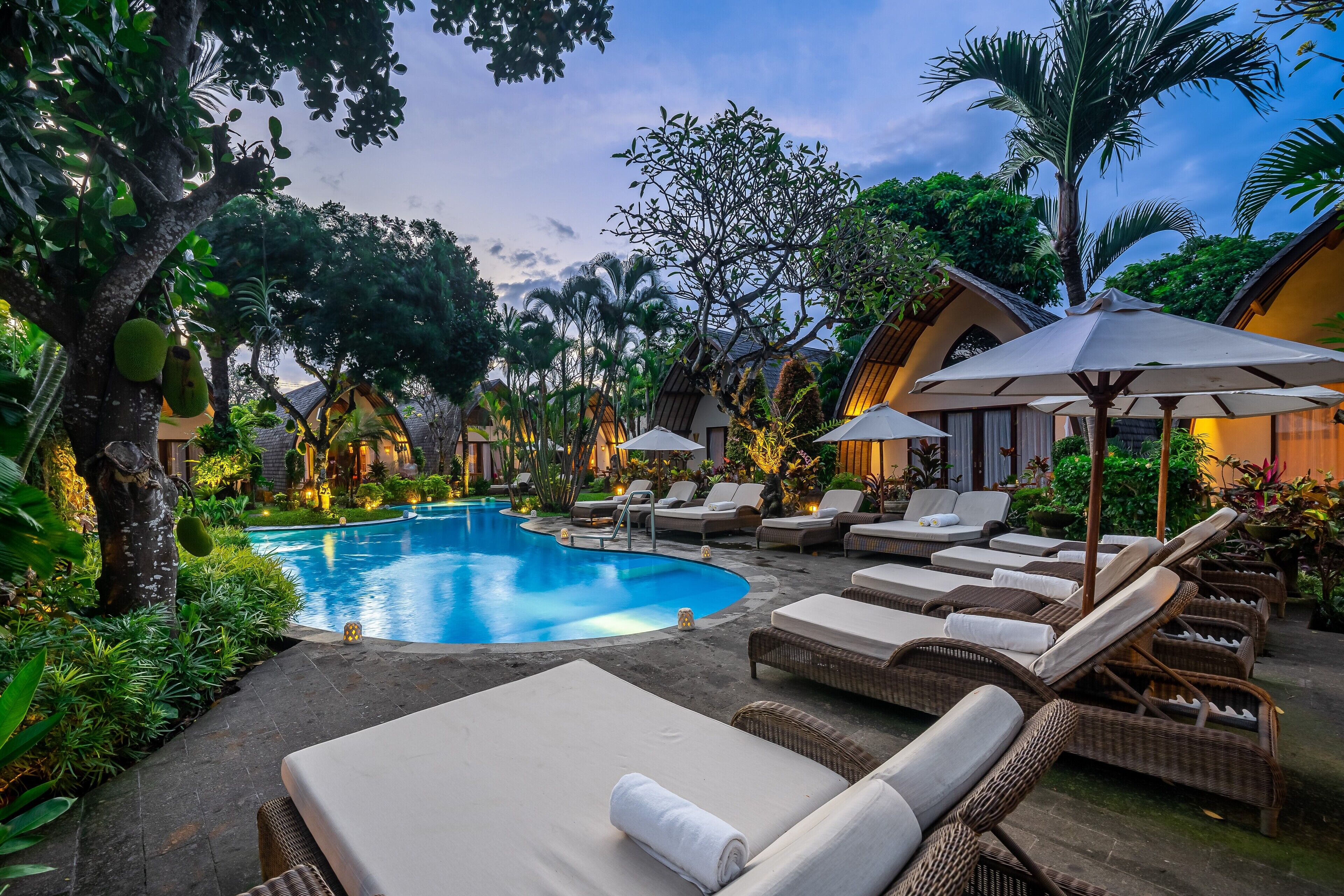 A poolside area with lounge chairs and umbrellas, surrounded by lush greenery and traditional architecture.