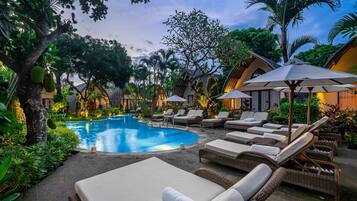 A poolside area with lounge chairs and umbrellas, surrounded by lush greenery and traditional architecture.