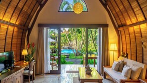 A spacious living area with a television, sofa, and dining table. A large window offers a view of a swimming pool and lush greenery. - Klumpu Bali Resort (Denpasar)