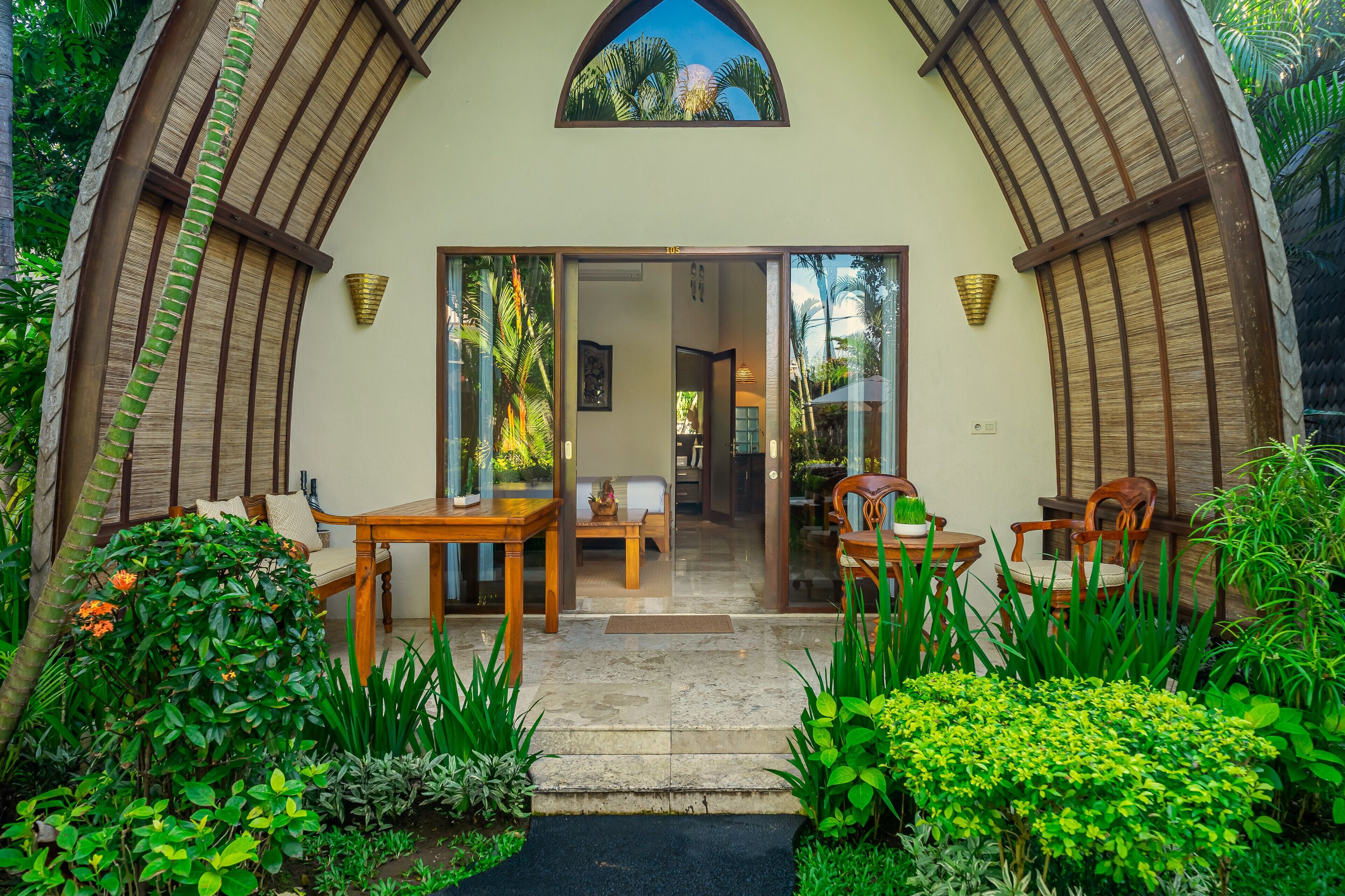 A villa with a covered entrance, a dining area with wooden furniture, and lush tropical plants.