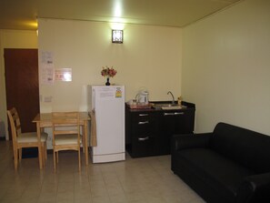 A compact living space with a dining table, chairs, a refrigerator, a black sofa, and a kitchenette area. - SK Muangthongthani Apartment (Pak Kret)