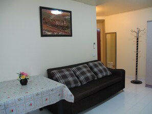 A living room with a dark sofa, a table with a floral tablecloth, and a framed picture on the wall. - SK Muangthongthani Apartment (Pak Kret)