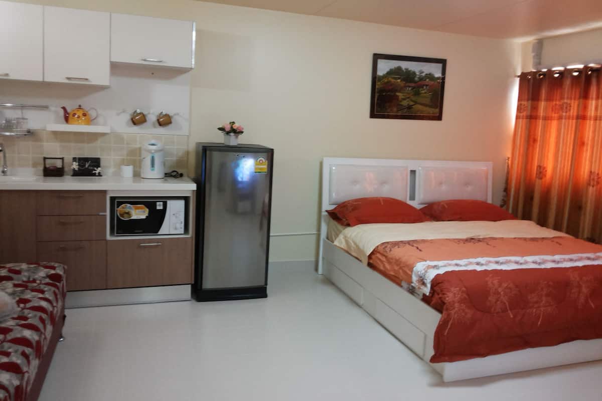 A compact hotel room with a kitchenette, a bed with red bedding, and a small window with curtains.
