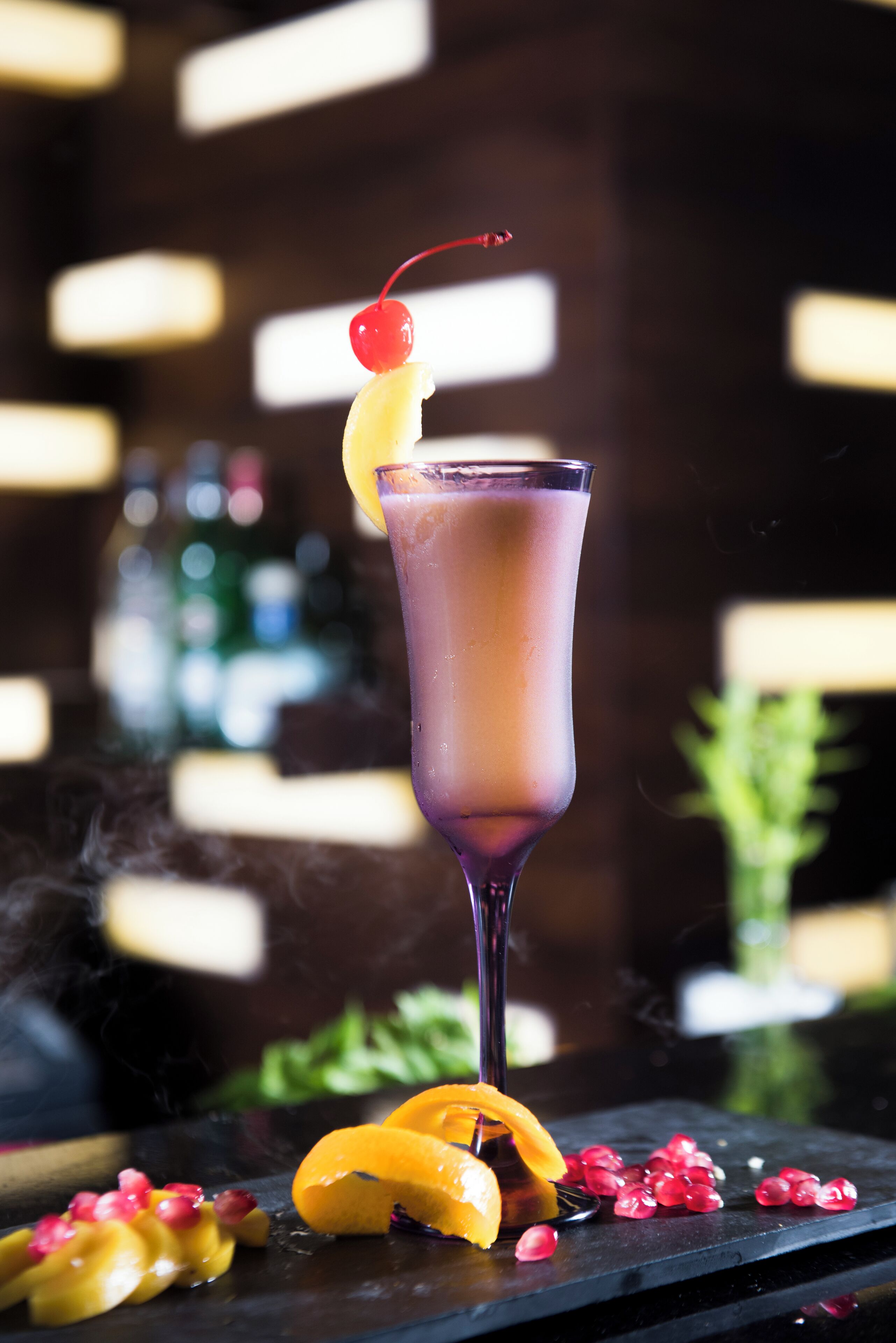 A cocktail with a cherry garnish, surrounded by pomegranate seeds and an orange slice on a dark surface.