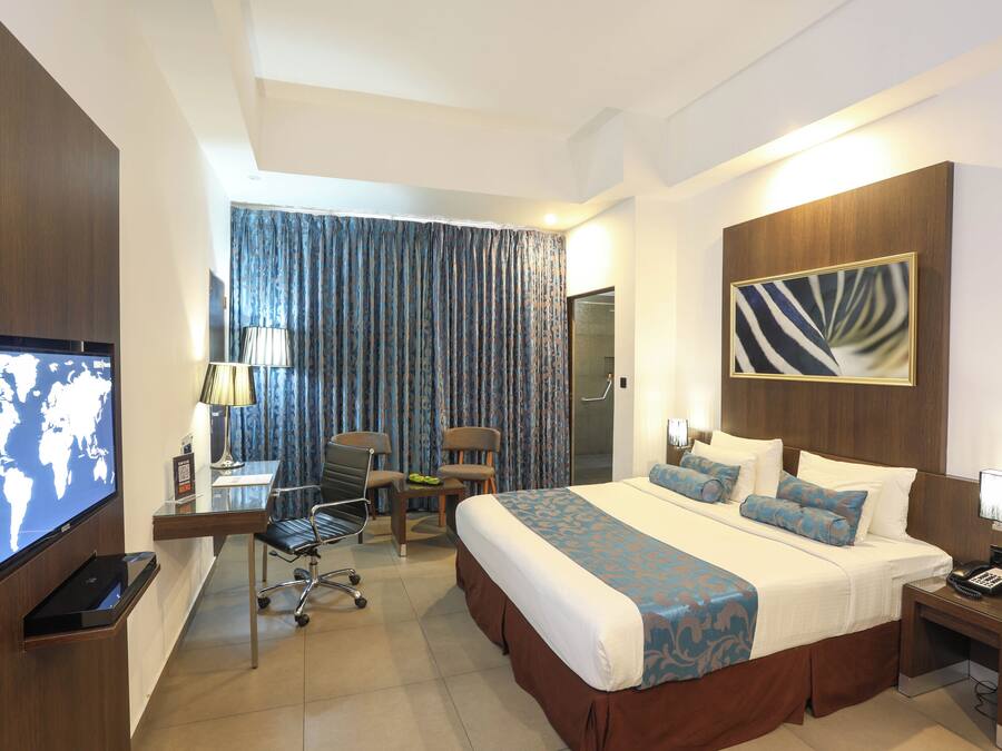 A hotel room with a large bed, a desk with a chair, a television, and a view of the outside through curtains.