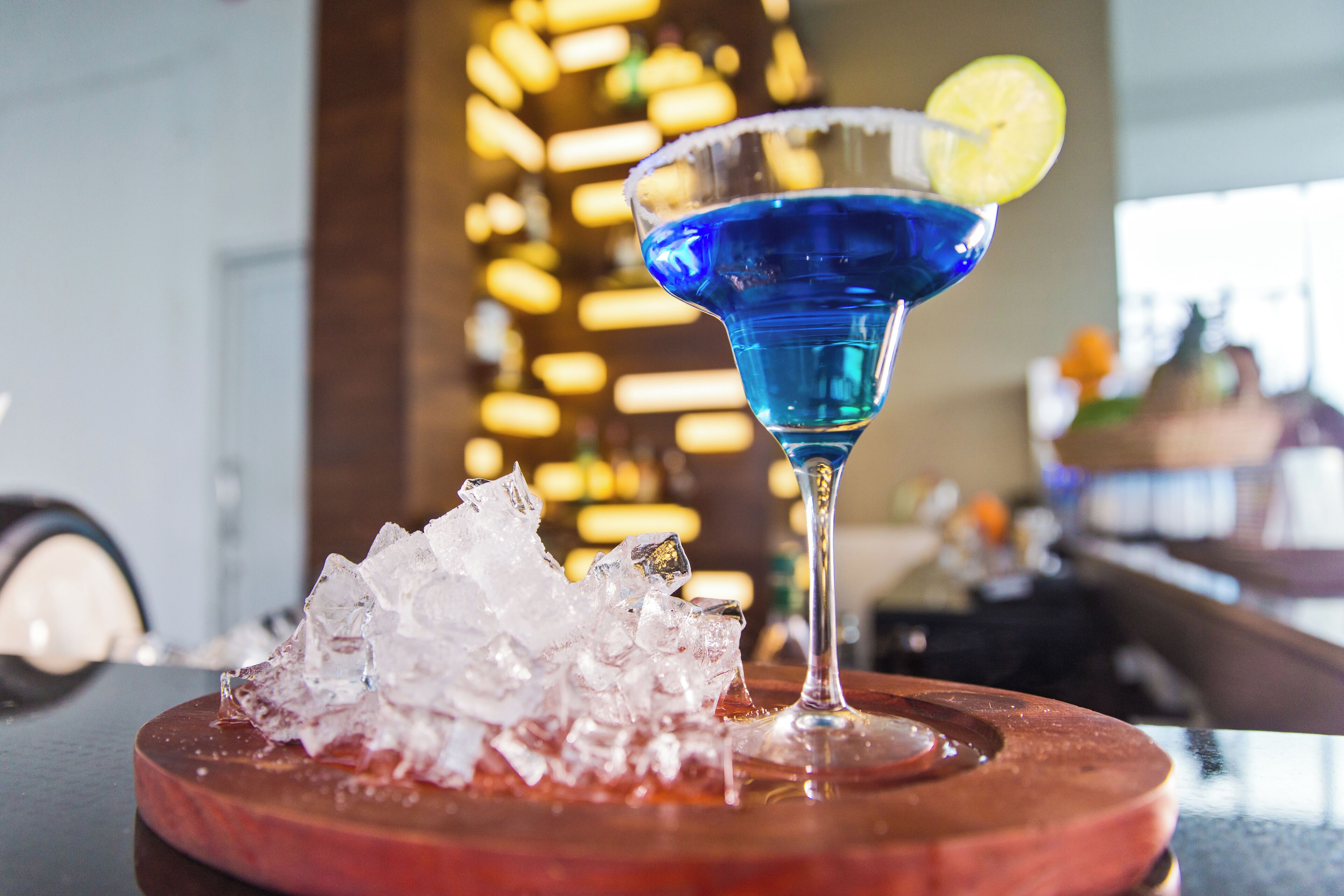 Cóctel azul con adorno de lima en una copa de martini, cubitos de hielo y un posavasos de madera.