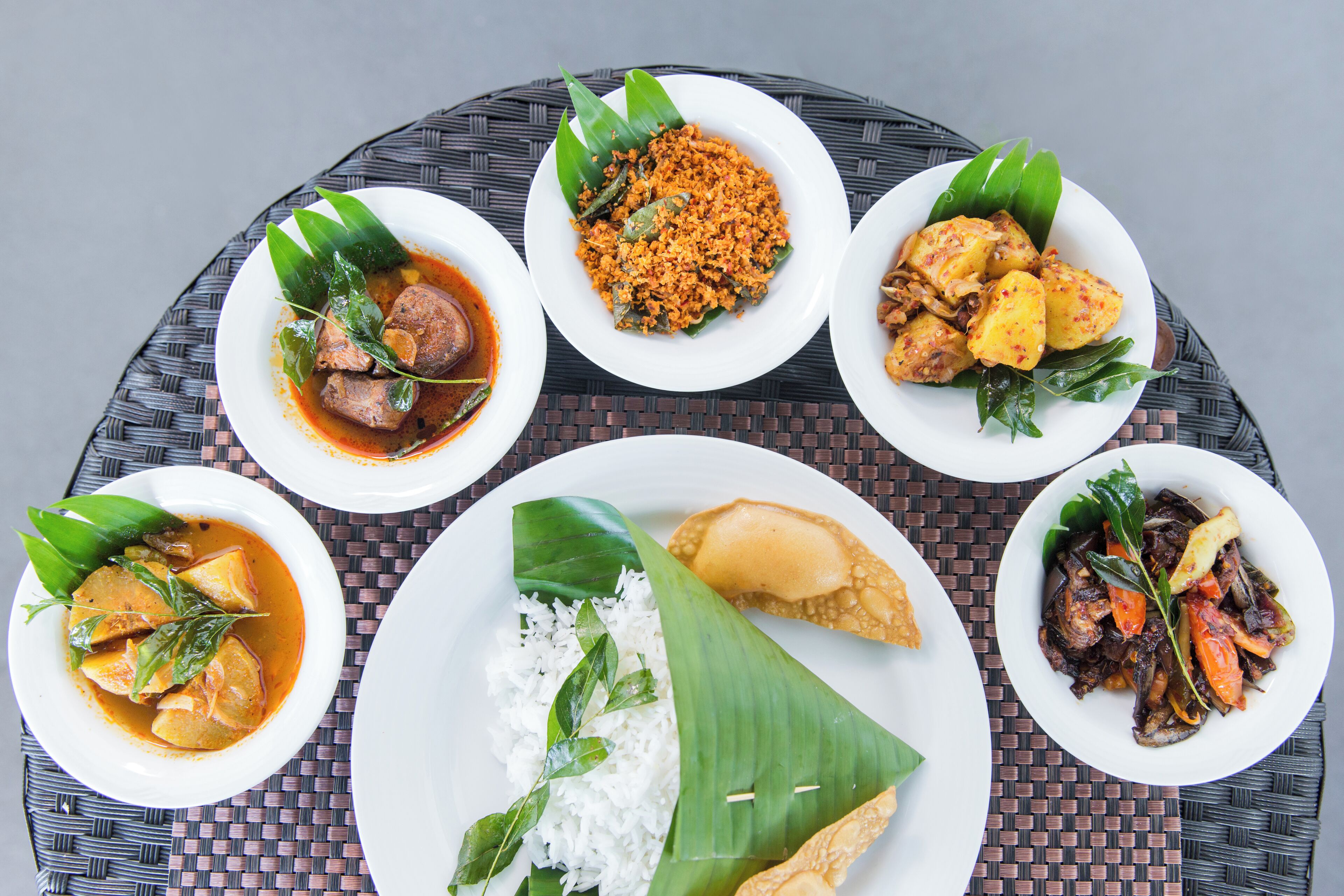 A table with various dishes, including rice, curry, and fried items, arranged on a woven tablecloth.