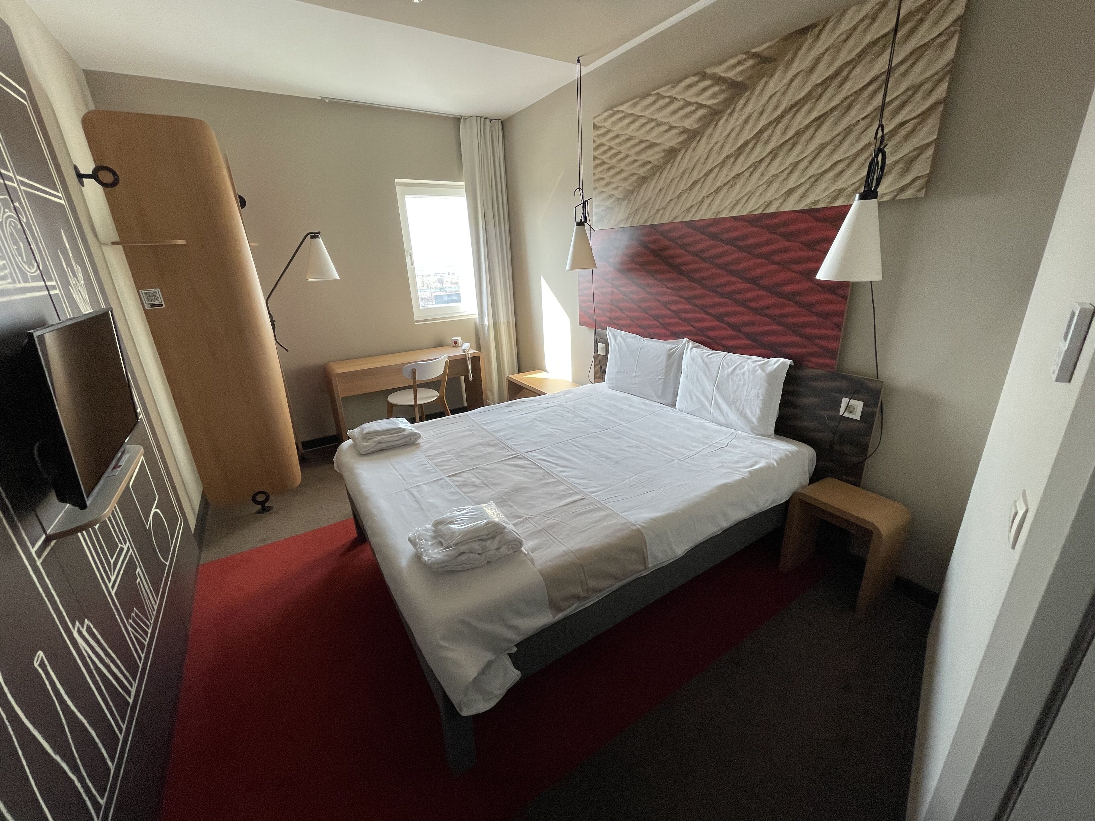 A hotel room with a bed, a desk, a chair, a TV, and a large window.