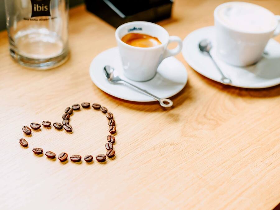 A table with two white cups of coffee, a spoon, and a heart-shaped arrangement of coffee beans.