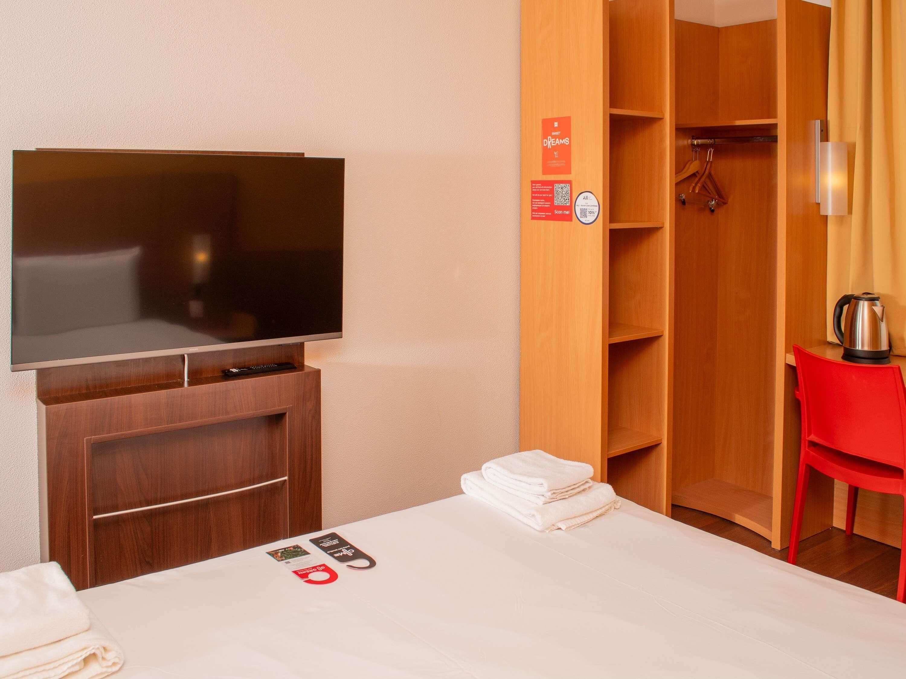 A hotel room with a flat-screen TV, a wooden wardrobe, a red chair, and a kettle on the table.
