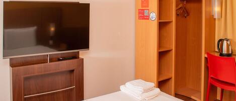 A hotel room with a flat-screen TV, a wooden wardrobe, a red chair, and a kettle on the table.
