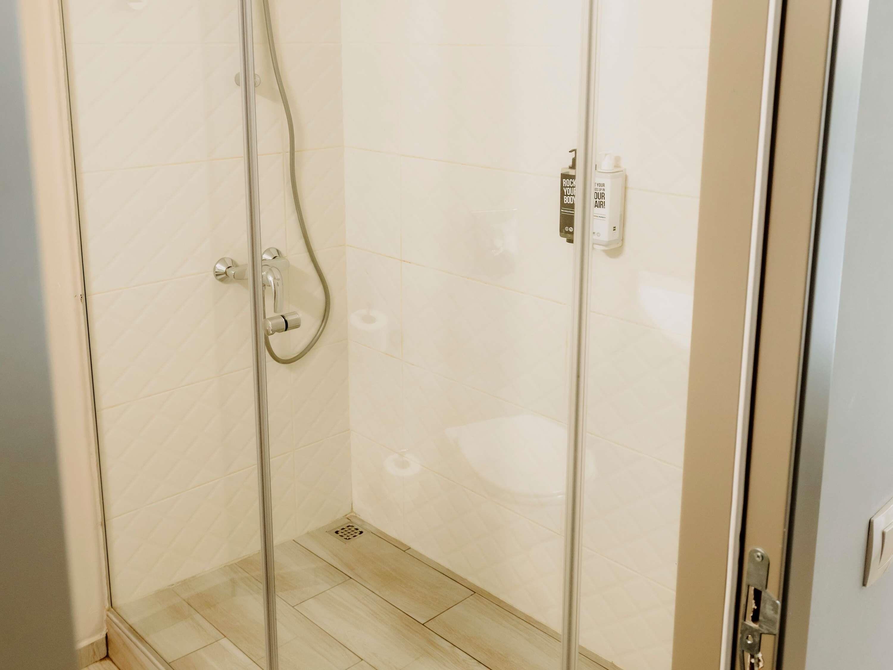 A glass-enclosed shower with a handheld showerhead, a wall-mounted soap dispenser, and a toilet paper holder on the wall.