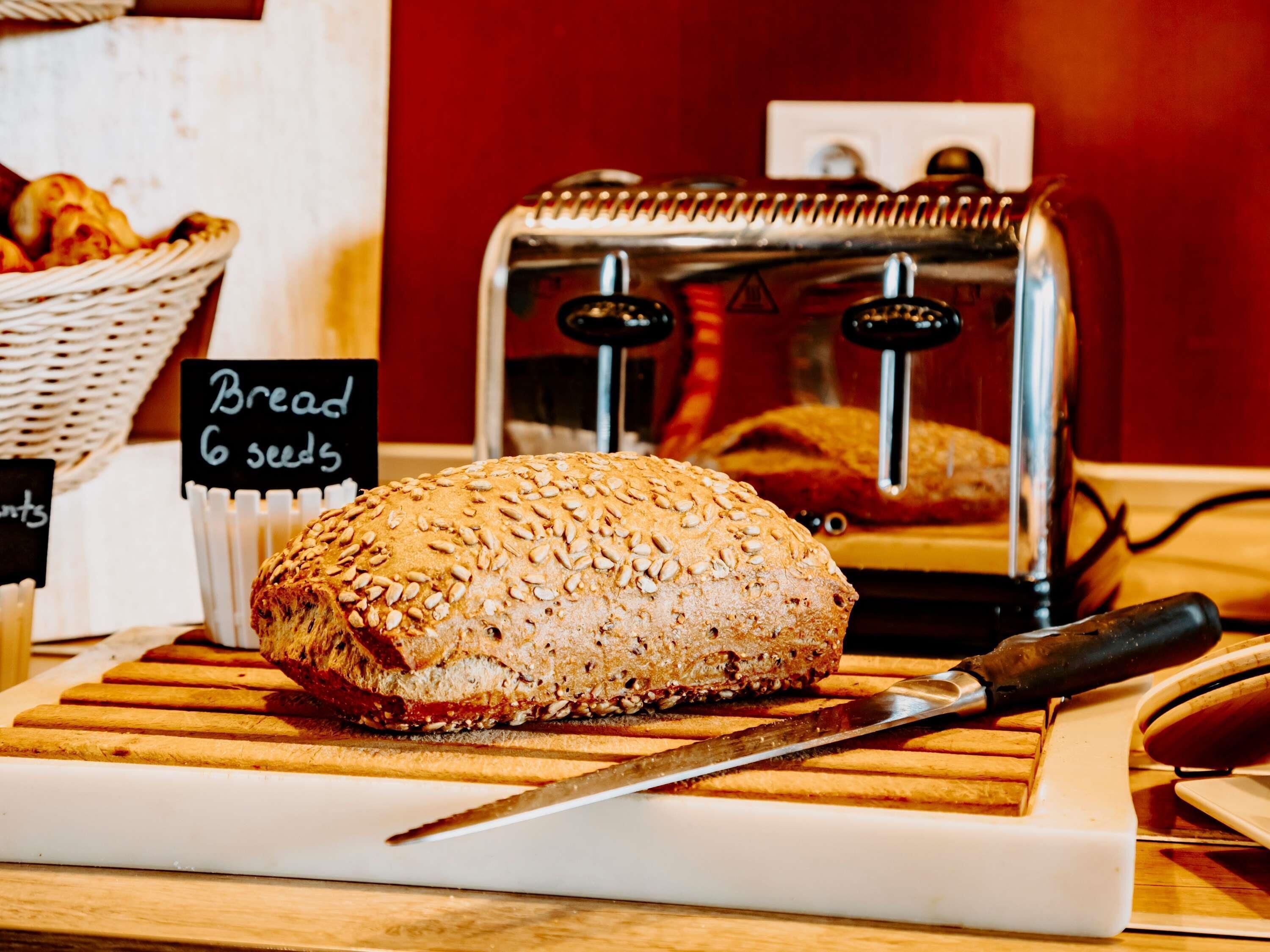 A loaf of bread with a sign indicating 'Bread 6 seeds' on a wooden cutting board, a knife, and a toaster in the background.