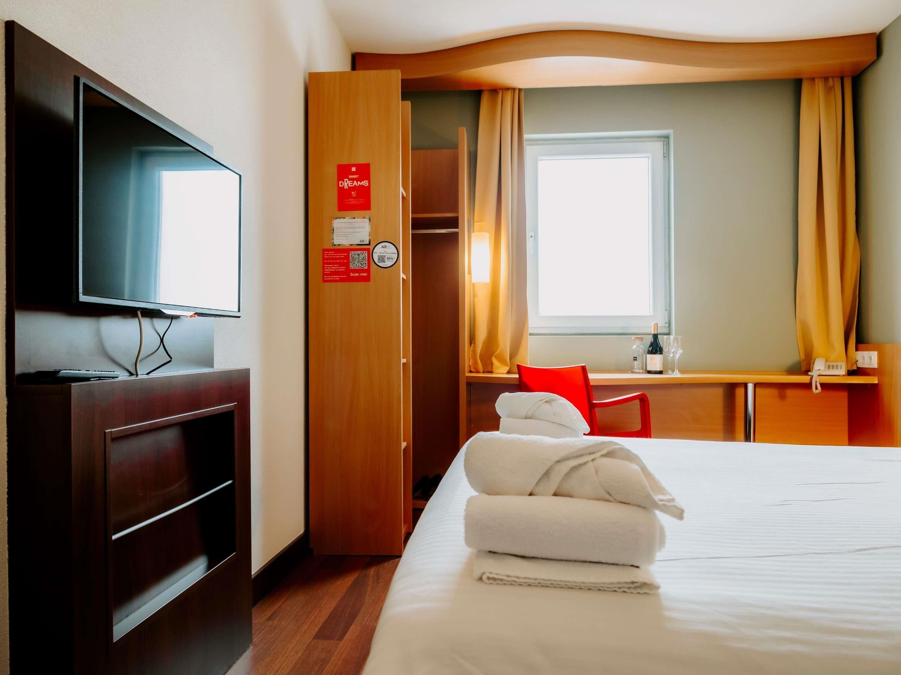 Une chambre d’hôtel avec un lit, une télévision, un bureau avec une chaise et une fenêtre avec des rideaux.