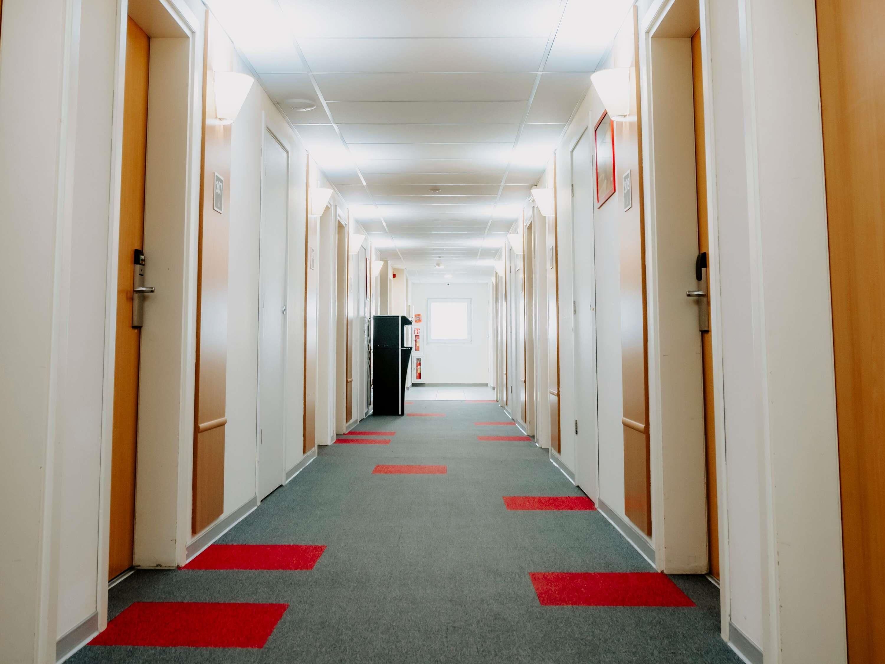 A long, narrow hotel corridor with doors on both sides, a grey carpet with red markings, and a bright ceiling.