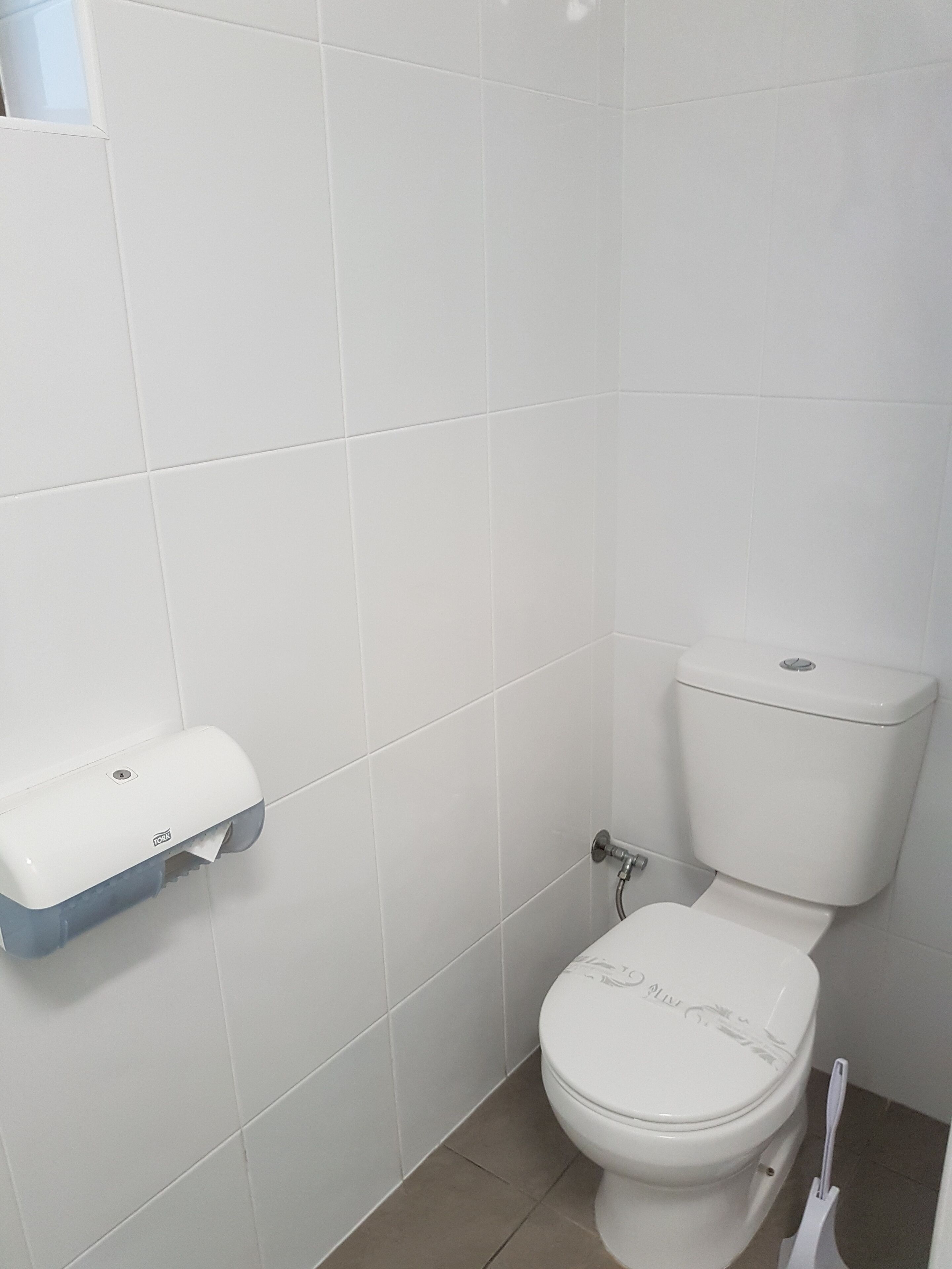 Een kleine, schone badkamer met witte tegels, een toilet, een papieren handdoekdispenser en een WC-borstel.