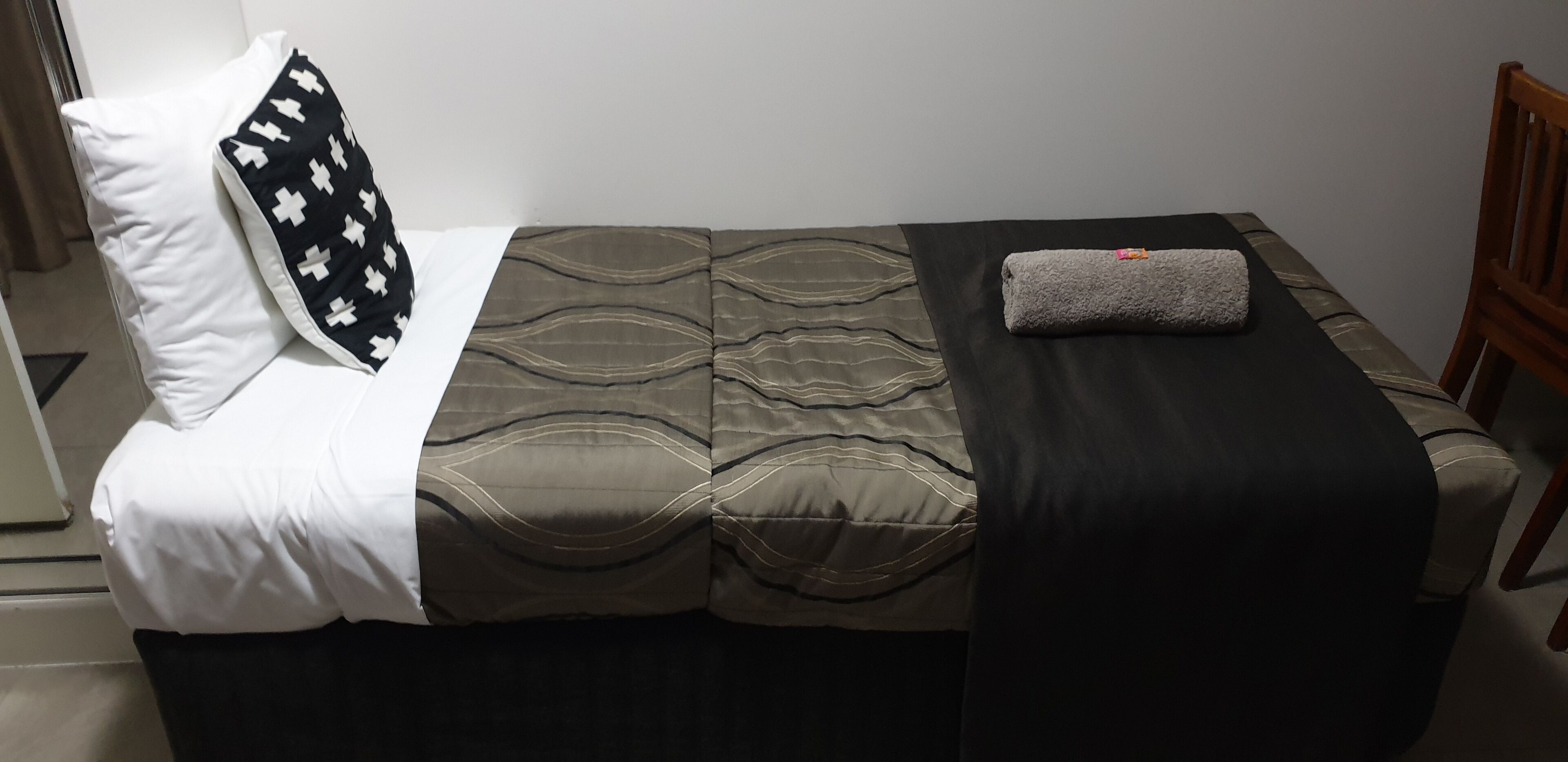 A single bed with a patterned bedspread, a black and white pillow, and a folded towel on the side.
